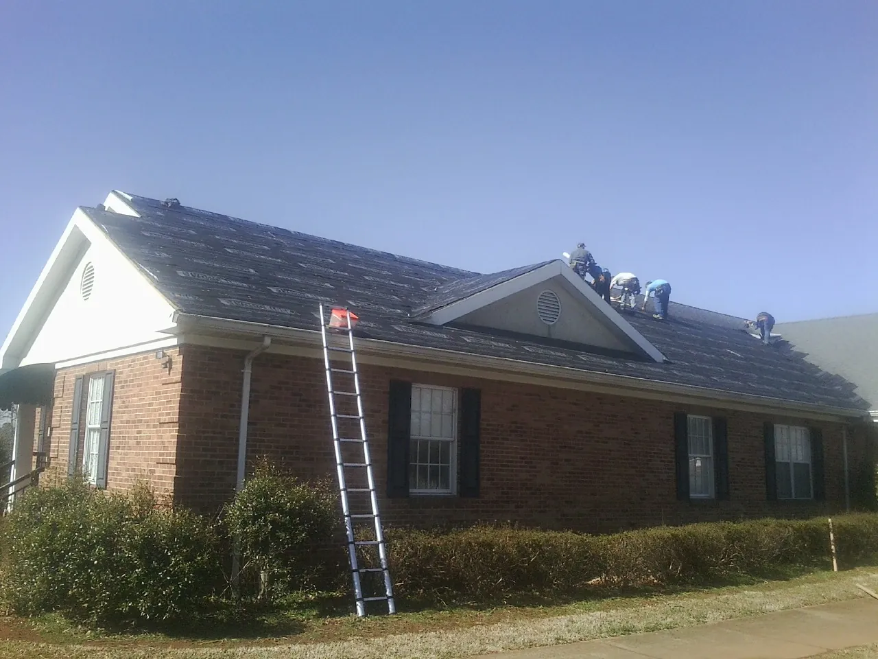 Crew tearing shingles off a brick ranch roof with a ladder set — Emergency Roof Repair work by our crew in Okarche