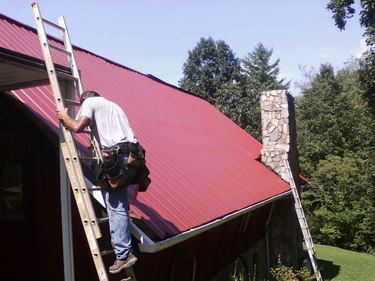 Roofer on a ladder beside a red metal roof and stone chimney — Soffit & Fascia Repair by Roofing Brothers in Okarche